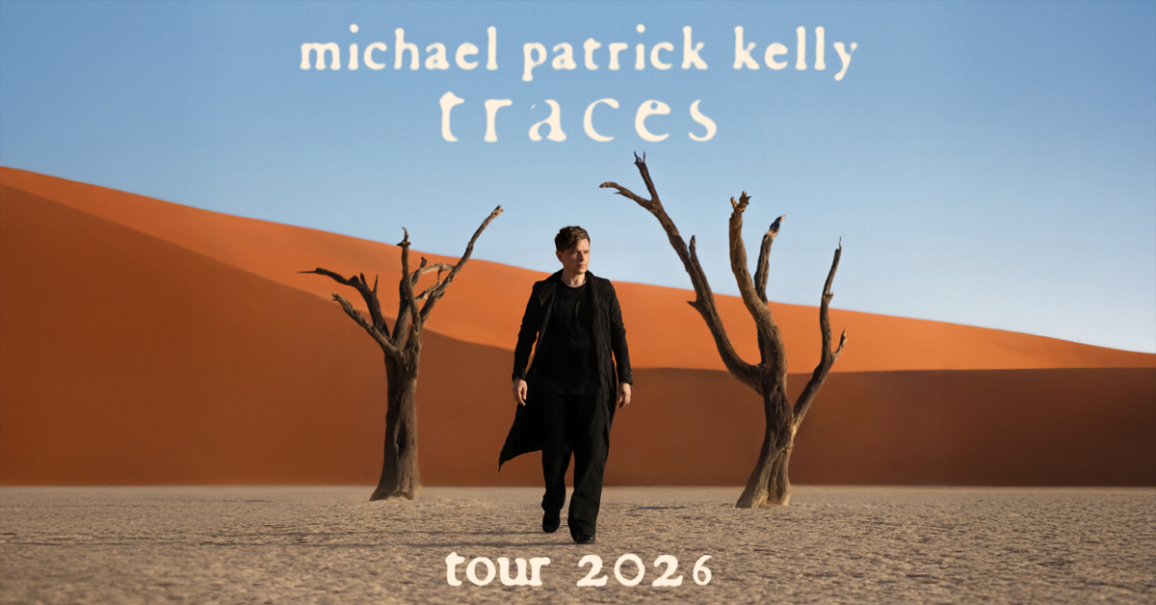 Ein Mann in schwarzer Kleidung steht in einer W&uuml;stenlandschaft mit kahlen B&auml;umen und rotem Sand. Der Himmel ist blau. Text: Michael Patrick Kelly - Traces Tour 2026., &copy; links im Bild