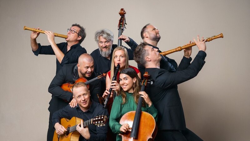Acht Musiker posieren mit verschiedenen Instrumenten, darunter Fl&ouml;ten, Gitarre und Cello, vor einem neutralen Hintergrund. Sie wirken fr&ouml;hlich und kreativ., &copy; Theaterhaus Stuttgart e.V.
