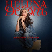 Eine Frau in einem Jeans-Outfit sitzt auf einem Holzkasten vor einem dunkelblauen Hintergrund. Der Text 'Helena Delphi' und 'Wanderer Tour 2026' ist zu sehen., &copy; links im Bild