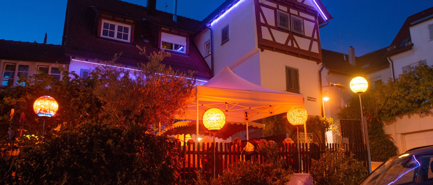 An illuminated half-timbered house with a white tent in the garden. Colorful lanterns and fairy lights create a cozy atmosphere at night., © Weingut Ruoff