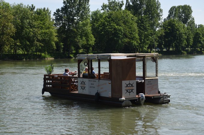 Ein rustikales Floßboot mit überdachtem Bereich fährt auf einem Fluss. Im Hintergrund sind grüne Bäume zu sehen., © Cool-Tours StattReisen Ein rustikales Floßboot mit überdachtem Bereich fährt auf einem Fluss. Im Hintergrund sind grüne Bäume zu sehen., © Cool-Tours StattReisen