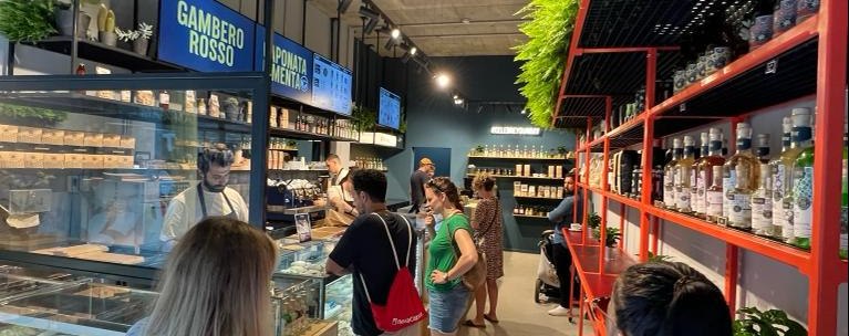 Lively delicatessen with plants on the ceiling, customers in a queue, shelves with products and a counter with food., &copy; CELEBRE