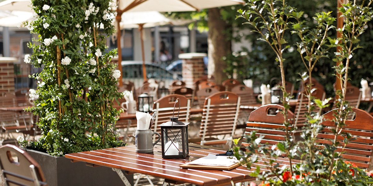 Ein gem&uuml;tlicher Biergarten mit Holztischen und -st&uuml;hlen, umgeben von gr&uuml;nen Pflanzen und Sonnenschirmen. Eine Laterne steht auf dem Tisch., &copy; Parkhotel Stuttgart Messe-Airport