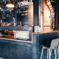 Modern bar with industrial design, high stools and wall art. Glasses are placed on the counter, in the background a table with drinks., © Kraftpaule Modern bar with industrial design, high stools and wall art. Glasses are placed on the counter, in the background a table with drinks., © Kraftpaule