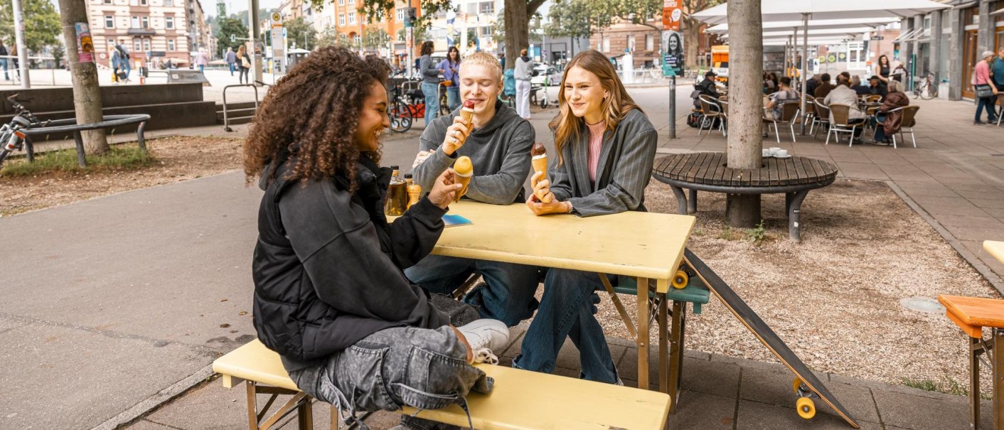 Drei Personen sitzen an einem gelben Tisch im Freien und essen Eis. Im Hintergrund sind Bäume, Fahrräder und ein Café zu sehen., © Stuttgart Marketing GmbH, Sarah Schmid Drei Personen sitzen an einem gelben Tisch im Freien und essen Eis. Im Hintergrund sind Bäume, Fahrräder und ein Café zu sehen., © Stuttgart Marketing GmbH, Sarah Schmid