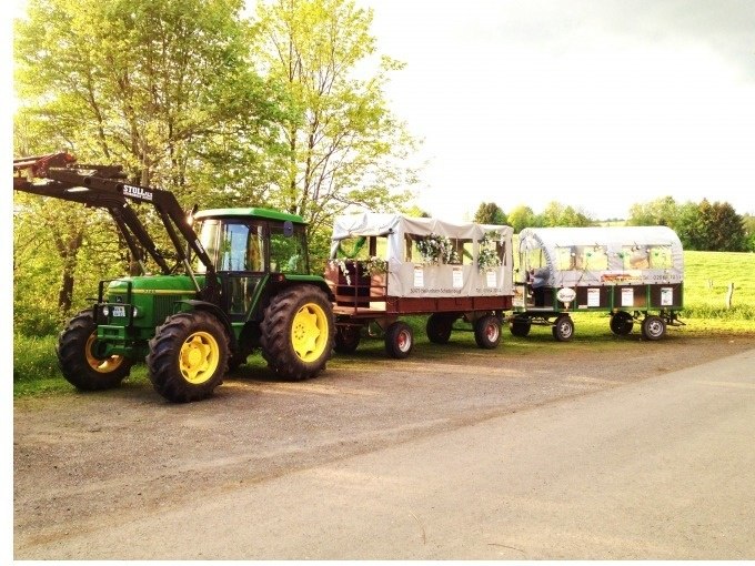 Ein gr&uuml;ner Traktor mit zwei Anh&auml;ngern steht auf einem Feldweg. Die Anh&auml;nger sind mit Planen bedeckt. Im Hintergrund sind B&auml;ume und Wiesen zu sehen., &copy; Cool-Tours StattReisen