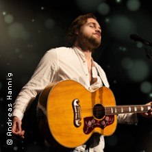 Ein Musiker mit weißem Hemd spielt Gitarre auf der Bühne. Der Hintergrund ist dunkel mit verschwommenen Lichtern., © links im Bild Ein Musiker mit weißem Hemd spielt Gitarre auf der Bühne. Der Hintergrund ist dunkel mit verschwommenen Lichtern., © links im Bild