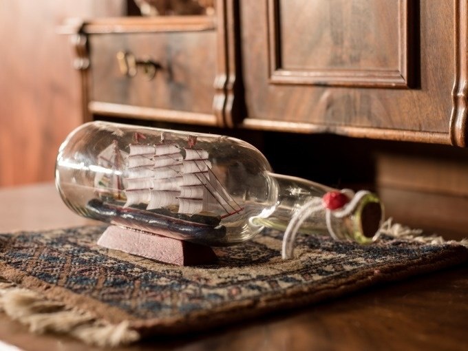 Ein Schiffsmodell in einer Flasche steht auf einem kleinen Teppich vor einem antiken Holzm&ouml;bel. Das Schiff hat wei&szlig;e Segel., &copy; ExitGames Stuttgart
