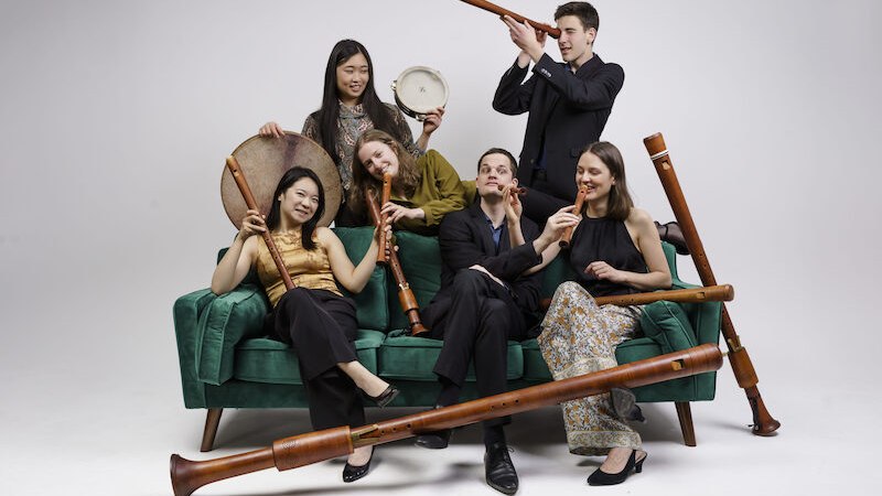 Sechs Musiker posieren mit Holzblasinstrumenten und einer Trommel auf einem grünen Sofa. Sie wirken fröhlich und kreativ., © Stiftsmusik Stuttgart