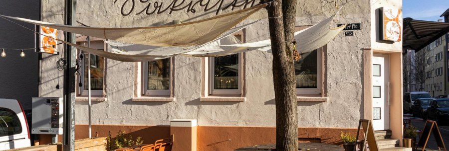 Ein Caf&eacute; mit Sonnensegeln und orangenen St&uuml;hlen im Au&szlig;enbereich. Die Fassade ist beige mit Schriftzug. Ein Baum steht vor dem Eingang., &copy; Stuttgart-Marketing GmbH, Sarah Schmid