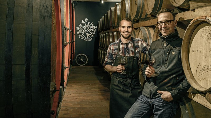 Zwei Männer in einem Weinkeller mit Holzfässern, halten Weingläser. Im Hintergrund ist ein Logo an der Wand., © Weingut Aldinger Zwei Männer in einem Weinkeller mit Holzfässern, halten Weingläser. Im Hintergrund ist ein Logo an der Wand., © Weingut Aldinger