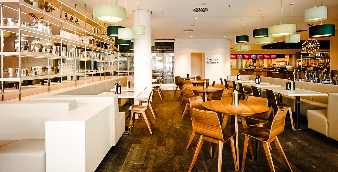 Modernes Restaurant mit Holztischen, weißen Bänken und Regalen voller Küchenutensilien. Grüne Lampen hängen von der Decke., © Breuninger