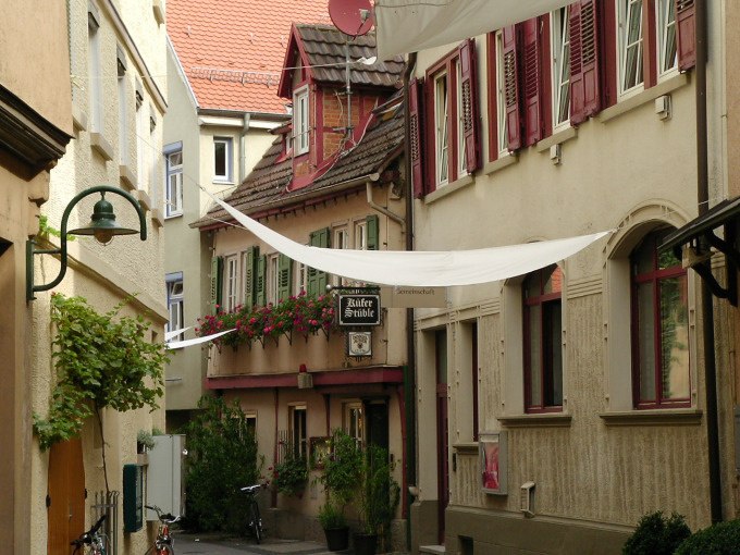 Narrow alley with historic buildings, red shutters and flowers. A sign points to a restaurant. White cloths stretch across the street., © Cool-Tours StattReisen Narrow alley with historic buildings, red shutters and flowers. A sign points to a restaurant. White cloths stretch across the street., © Cool-Tours StattReisen