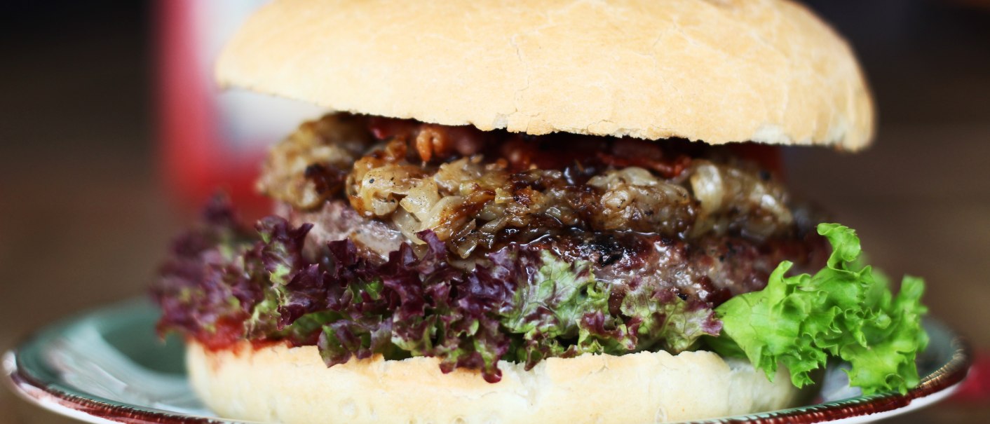 Ein saftiger Burger mit Salat und Fleischpatty auf einem Teller, im Hintergrund unscharf., © SMG