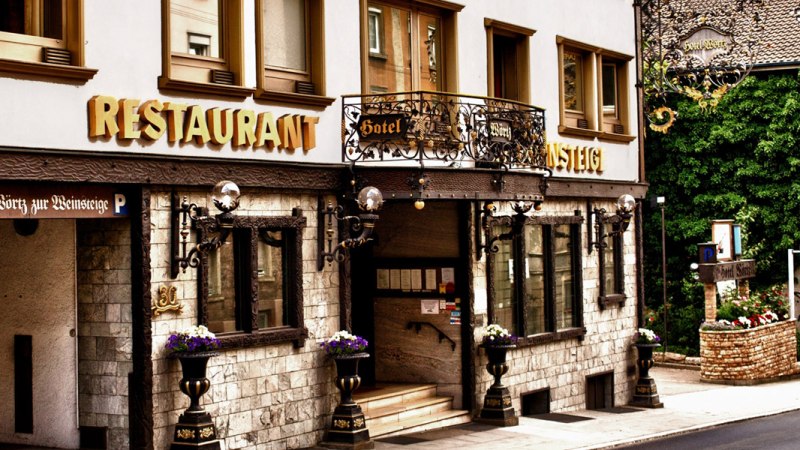 Eingang des Hotels Zur Weinsteige mit dekorativen Elementen, großen Fenstern und Restaurantbeschilderung. Blumen schmücken den Eingangsbereich., © Hotel Zur Weinsteige