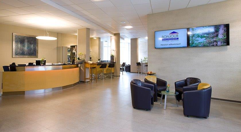 A modern lobby area with a reception desk, bar stools, and a cozy seating area. Two screens on the wall display welcome messages., &copy; bonjour Hotelbetriebs GmbH