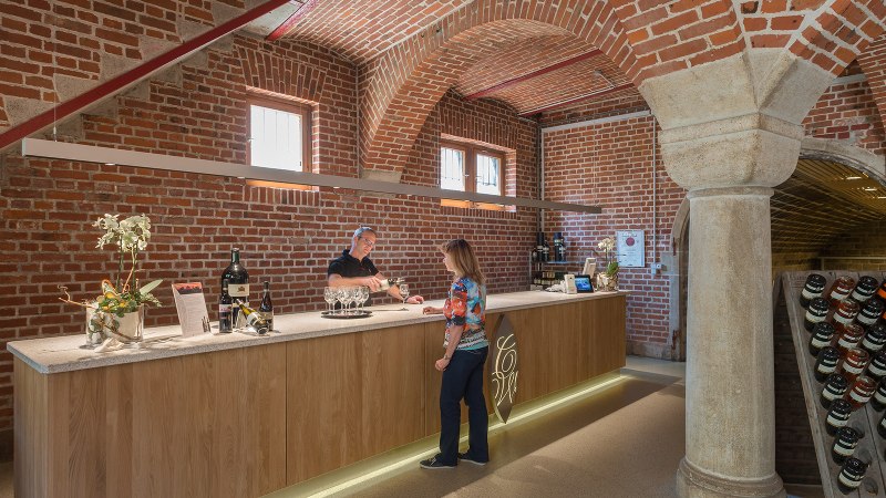 Eine Theke in einer Weinkellerei mit Backsteinwänden. Ein Mann schenkt einer Frau Wein ein. Weinflaschen sind auf der Theke ausgestellt., © Collegium Wirtemberg Eine Theke in einer Weinkellerei mit Backsteinwänden. Ein Mann schenkt einer Frau Wein ein. Weinflaschen sind auf der Theke ausgestellt., © Collegium Wirtemberg