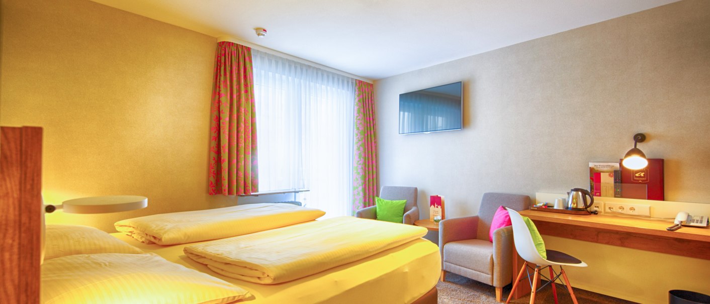 Ein gemütliches Hotelzimmer mit Doppelbett, Schreibtisch, Fernseher und zwei Sesseln. Helle Vorhänge und bunte Kissen sorgen für eine einladende Atmosphäre., © TOMAS Ein gemütliches Hotelzimmer mit Doppelbett, Schreibtisch, Fernseher und zwei Sesseln. Helle Vorhänge und bunte Kissen sorgen für eine einladende Atmosphäre., © TOMAS