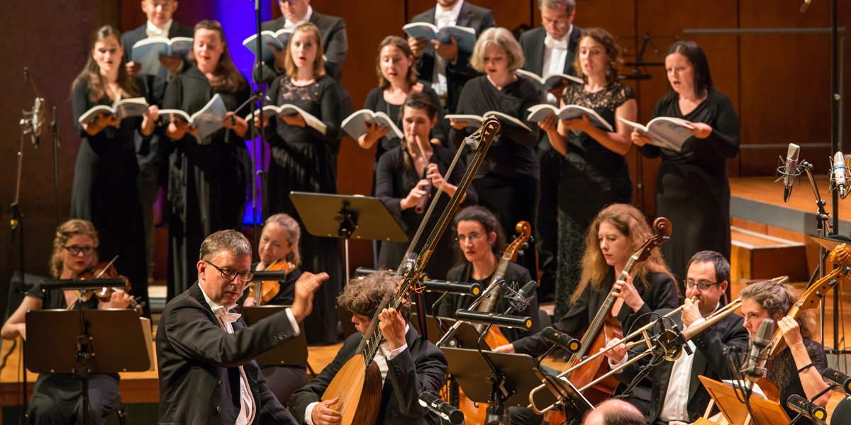 Ein Chor und Orchester in formeller Kleidung führen ein klassisches Musikstück auf. Der Dirigent leitet die Musiker, die konzentriert spielen und singen., © Holger Schneider Ein Chor und Orchester in formeller Kleidung führen ein klassisches Musikstück auf. Der Dirigent leitet die Musiker, die konzentriert spielen und singen., © Holger Schneider