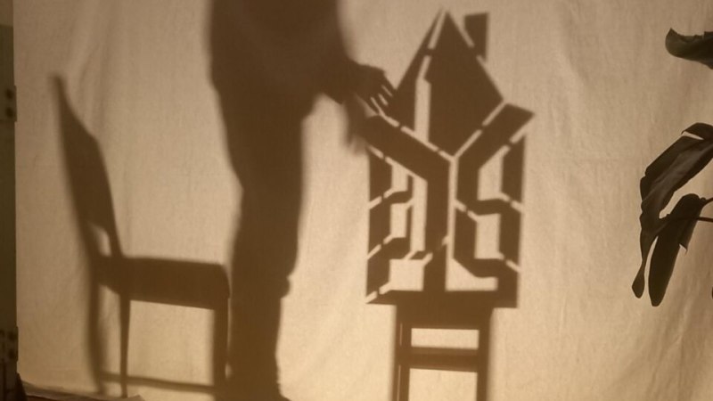 Ein Schattenspiel zeigt die Silhouette eines Menschen, der ein Haus auf einem Stuhl berührt. Die Szene ist auf eine Leinwand projiziert., © Esther Falk Ein Schattenspiel zeigt die Silhouette eines Menschen, der ein Haus auf einem Stuhl berührt. Die Szene ist auf eine Leinwand projiziert., © Esther Falk