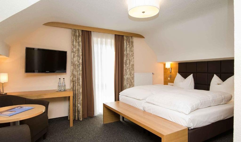 Modern double room with large bed, flat screen TV, seating area and table. Bright walls, curtains and carpeted floor., © Nödinger Gastro GmbH