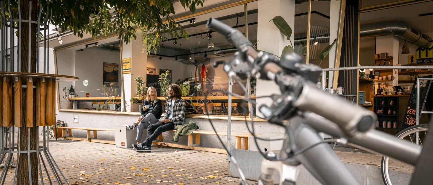 Zwei Personen sitzen lachend vor einem Café auf einer Bank. Im Vordergrund ist ein Fahrradlenker zu sehen. Herbstliche Blätter liegen auf dem Boden., © SMG Stuttgart Marketing GmbH - Sarah Schmid Zwei Personen sitzen lachend vor einem Café auf einer Bank. Im Vordergrund ist ein Fahrradlenker zu sehen. Herbstliche Blätter liegen auf dem Boden., © SMG Stuttgart Marketing GmbH - Sarah Schmid