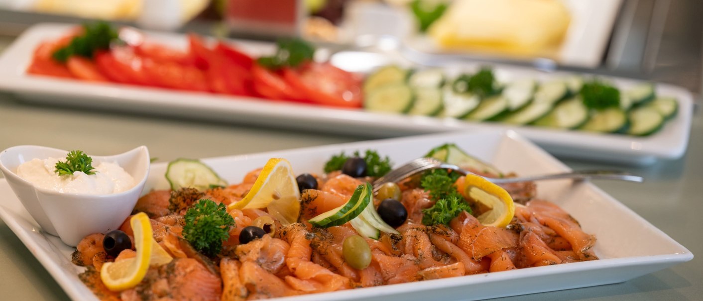 Geräucherter Lachs mit Zitronenscheiben und Oliven auf einem Teller, daneben eine Schale mit Sahne. Im Hintergrund sind Gemüseplatten zu sehen., © Erikson Hotel Geräucherter Lachs mit Zitronenscheiben und Oliven auf einem Teller, daneben eine Schale mit Sahne. Im Hintergrund sind Gemüseplatten zu sehen., © Erikson Hotel