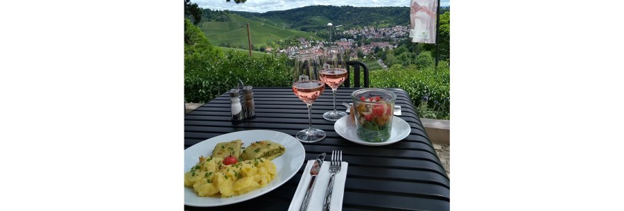 A table with two plates of food and two glasses of rosé wine. Vineyards and a village in the background., © 1819 Bistro am Wirtemberg A table with two plates of food and two glasses of rosé wine. Vineyards and a village in the background., © 1819 Bistro am Wirtemberg