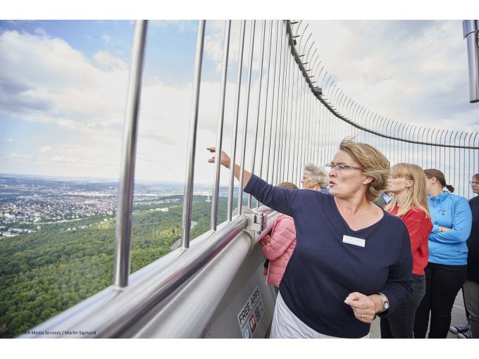 Eine Frau zeigt von einem Aussichtsturm auf die Landschaft, w&auml;hrend mehrere Personen zuschauen., &copy; SWR Media Services GmbH / Fernsehturm Stuttgart