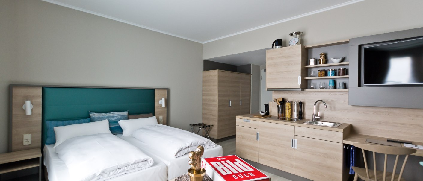 Modernes Apartment mit Doppelbett, K&uuml;chenzeile, Tisch und Stuhl. Ein Buch liegt auf dem Tisch, daneben eine goldene Figur., &copy; Arthotel Ana / elaya