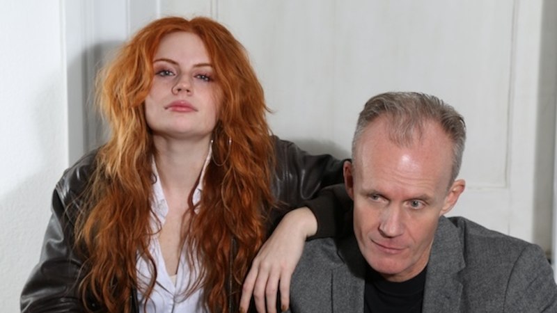 Eine Frau mit roten Haaren und ein Mann in einem grauen Blazer sitzen nebeneinander vor einer wei&szlig;en Wand. Sie wirkt entspannt, er nachdenklich., &copy; Renitenztheater Stuttgart e.V.