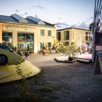 Evening event at the Kunstverein Wagenhalle with an art installation of a yellow car and people sitting in the square watching a performance., © Atelier Oradoro Evening event at the Kunstverein Wagenhalle with an art installation of a yellow car and people sitting in the square watching a performance., © Atelier Oradoro