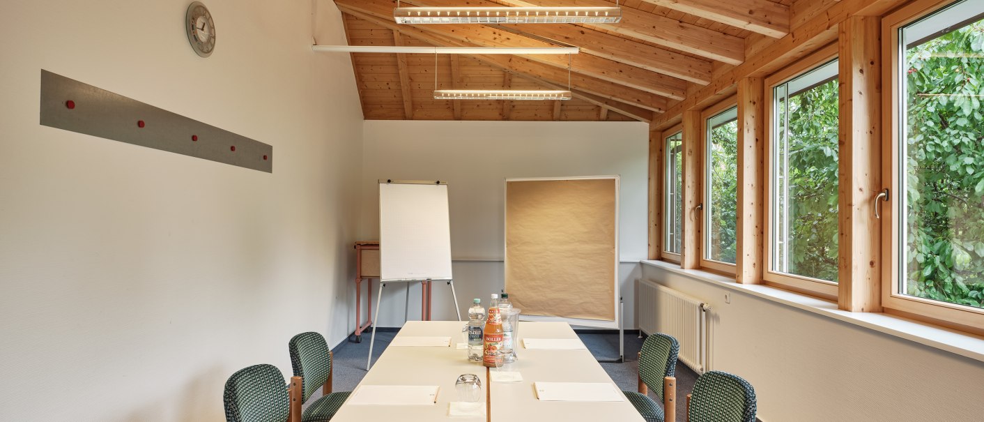 A bright seminar room with a wooden ceiling, a table, and chairs. A flip chart is set up. Large windows offer a view of the greenery outside., © Badhotel Restaurant Stauferland A bright seminar room with a wooden ceiling, a table, and chairs. A flip chart is set up. Large windows offer a view of the greenery outside., © Badhotel Restaurant Stauferland