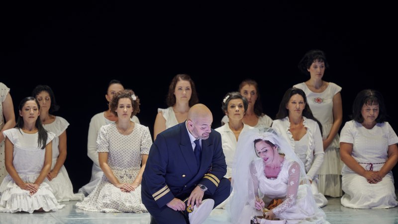 Ein Mann in Uniform kniet neben einer Frau im Brautkleid. Um sie herum sitzen Frauen in wei&szlig;en Kleidern. Die Szene wirkt dramatisch und emotional., &copy; Martin Sigmund