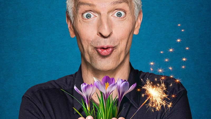 Ein Mann mit grauen Haaren hält Krokusse und eine Wunderkerze in den Händen vor blauem Hintergrund., © Renitenztheater Stuttgart e.V.
