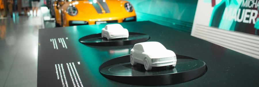 Model cars on a table in the Porsche Museum, an orange Porsche and visitors in the background. On the right, a poster with the name Michael Mauer., © Stuttgart Marketing GmbH, Sarah Schmid