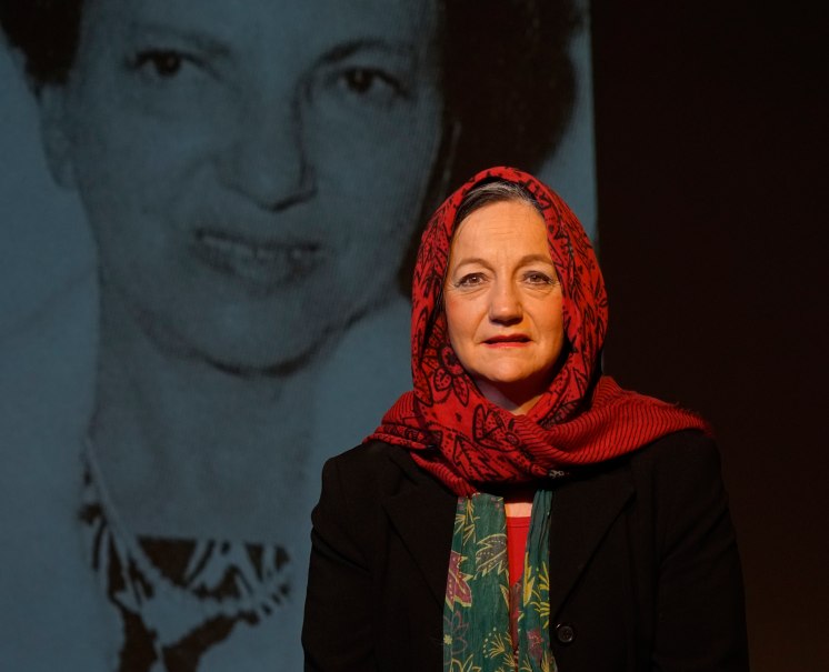 Eine Frau mit rotem Kopftuch steht vor einem großen Schwarz-Weiß-Foto einer anderen Frau. Sie trägt einen schwarzen Blazer und schaut direkt in die Kamera., © ©Katja Ritter Eine Frau mit rotem Kopftuch steht vor einem großen Schwarz-Weiß-Foto einer anderen Frau. Sie trägt einen schwarzen Blazer und schaut direkt in die Kamera., © ©Katja Ritter