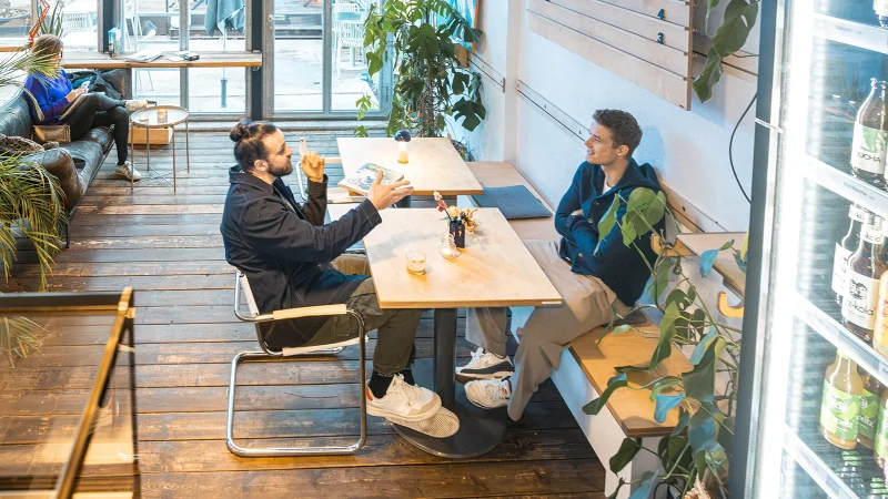 Zwei M&auml;nner unterhalten sich in einem gem&uuml;tlichen Caf&eacute;; im Hintergrund sitzt eine Frau auf einem Sofa. Rechts ist ein K&uuml;hlschrank mit Getr&auml;nken zu sehen., &copy; SMG, Sarah Schmid