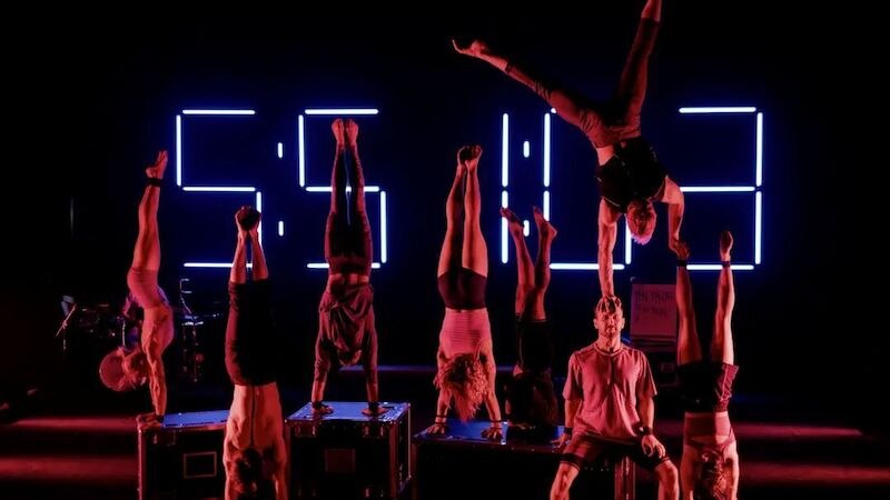 Acht Akrobaten machen Handstände auf einer Bühne. Im Hintergrund leuchten große Zahlen in Blau. Die Szene ist dramatisch beleuchtet., © Theaterhaus Stuttgart e.V. Acht Akrobaten machen Handstände auf einer Bühne. Im Hintergrund leuchten große Zahlen in Blau. Die Szene ist dramatisch beleuchtet., © Theaterhaus Stuttgart e.V.
