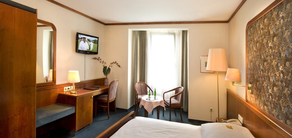 Gem&uuml;tliches Hotelzimmer mit Doppelbett, Fernseher, Schreibtisch und Sitzbereich vor einem Fenster. Holzm&ouml;bel und warme Beleuchtung dominieren den Raum., &copy; Hotel am Schelztor