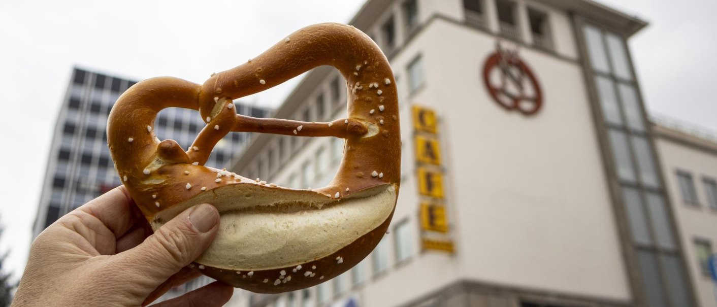 Eine Hand hält eine Brezel vor einem Gebäude mit dem Schriftzug 'Café' und einem Brezel-Logo., © Stuttgart-Marketing GmbH, Sarah Schmid Eine Hand hält eine Brezel vor einem Gebäude mit dem Schriftzug 'Café' und einem Brezel-Logo., © Stuttgart-Marketing GmbH, Sarah Schmid