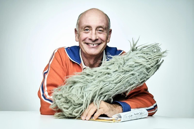 Ein Mann in einer orangefarbenen Jacke lächelt und hält ein graues, flauschiges Objekt. Vor ihm liegt ein Notizbuch., © Theaterhaus Stuttgart e.V.