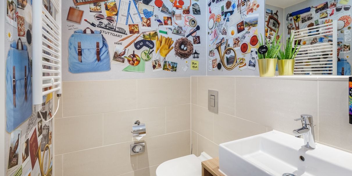 Modernes Badezimmer mit Collage-Wand, weißen Fliesen, Waschbecken und Toilette. Pflanzen in goldenen Töpfen als Dekoration., © Novum Hospitality