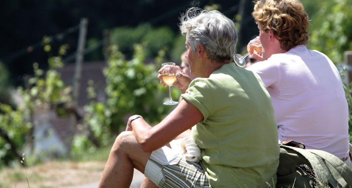 Two people sit on the grass and enjoy a glass of wine on a wine trail. Vines can be seen in the background., © Stuttgart-Marketing GmbH Two people sit on the grass and enjoy a glass of wine on a wine trail. Vines can be seen in the background., © Stuttgart-Marketing GmbH