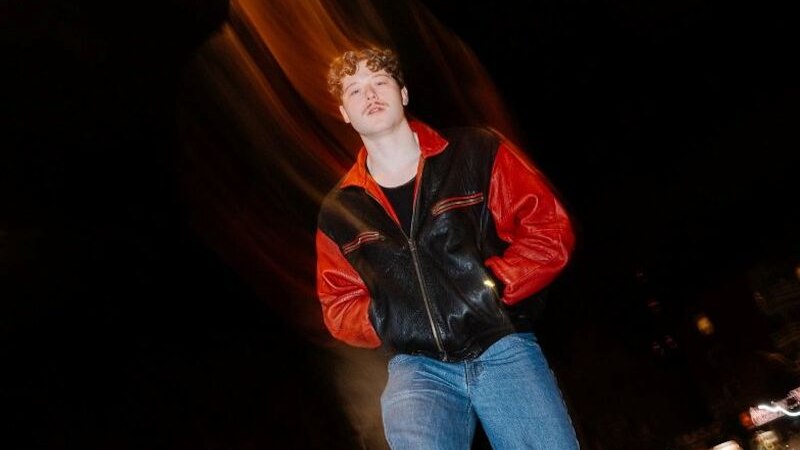 A man in a black and red leather jacket stands on a street at night. The background is blurred, suggesting dynamic movement., &copy; Marvin Hoffmann