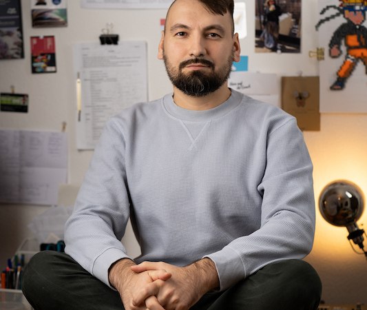 Person sitzt im Schneidersitz auf einem Tisch, umgeben von Zeitungsausschnitten und Bildern an der Wand., © Rosenau Kultur e.V.