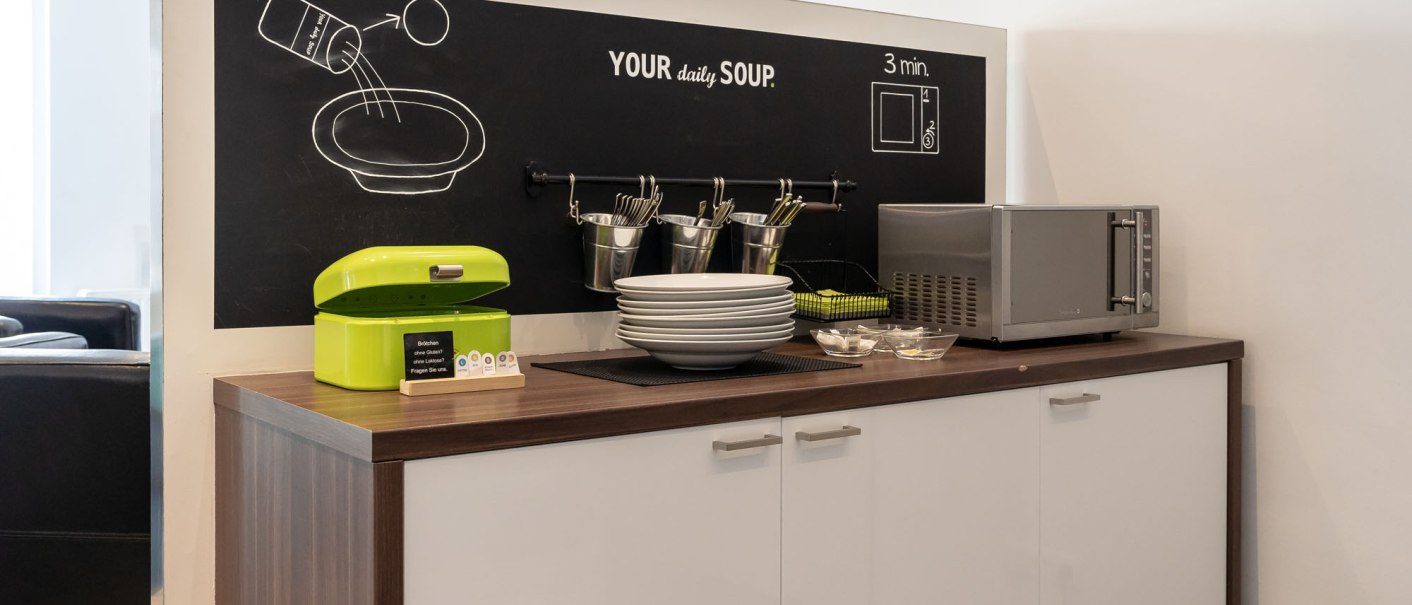 Küchenzeile mit Mikrowelle, grüner Brotdose und Geschirr. An der Wand eine Tafel mit Suppenillustration und der Aufschrift 'Your daily soup'., © a2 HOTELS Küchenzeile mit Mikrowelle, grüner Brotdose und Geschirr. An der Wand eine Tafel mit Suppenillustration und der Aufschrift 'Your daily soup'., © a2 HOTELS
