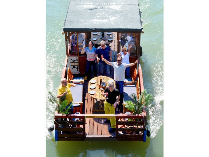 A group of people are standing on a raft boat with a table and chairs as it sails down a river. They appear cheerful and relaxed., © Cool-Tours StattReisen A group of people are standing on a raft boat with a table and chairs as it sails down a river. They appear cheerful and relaxed., © Cool-Tours StattReisen
