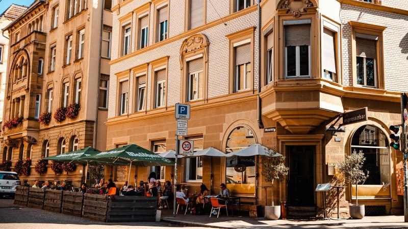 Straßencafé mit Sonnenschirmen vor einem historischen Gebäude. Menschen sitzen draußen, genießen das Wetter. Verkehrsschilder und eine Vespa sind sichtbar., © Stuttgart Marketing GmbH, Sarah Schmid