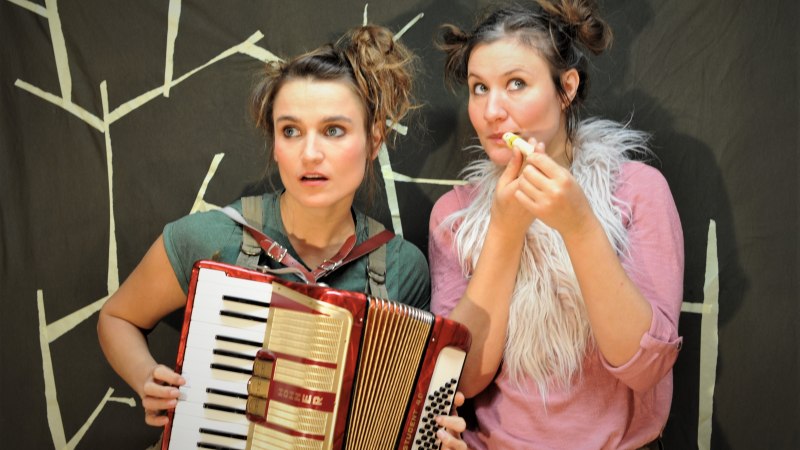 Zwei Frauen vor einem dunklen Hintergrund mit weißen Linien. Eine spielt Akkordeon, die andere trägt eine flauschige Weste und hält einen Lippenpflegestift., © Anetta Dick Zwei Frauen vor einem dunklen Hintergrund mit weißen Linien. Eine spielt Akkordeon, die andere trägt eine flauschige Weste und hält einen Lippenpflegestift., © Anetta Dick